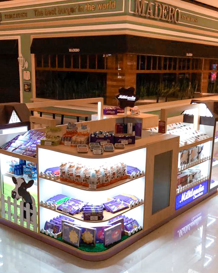 Ponto de venda da Milka. A imagem mostra toda a bancada pegando a ponta dela, que mostra dois lados simultaneamente além do topo. Na bancada tem alguns produtos expostos, além de mostrar placas de sinalização com escritos de "milklandia", além de várias sinalizações em MDF.